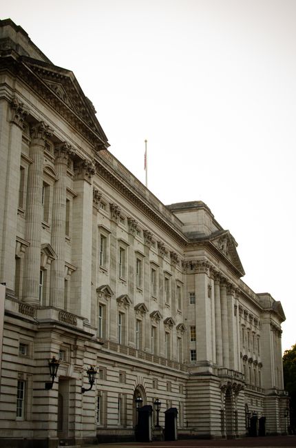 A large, historic palace building situated behind a lush, green park with a tranquil lake in the foreground. The palace's ornate, classical architecture includes detailed stonework, tall windows, and